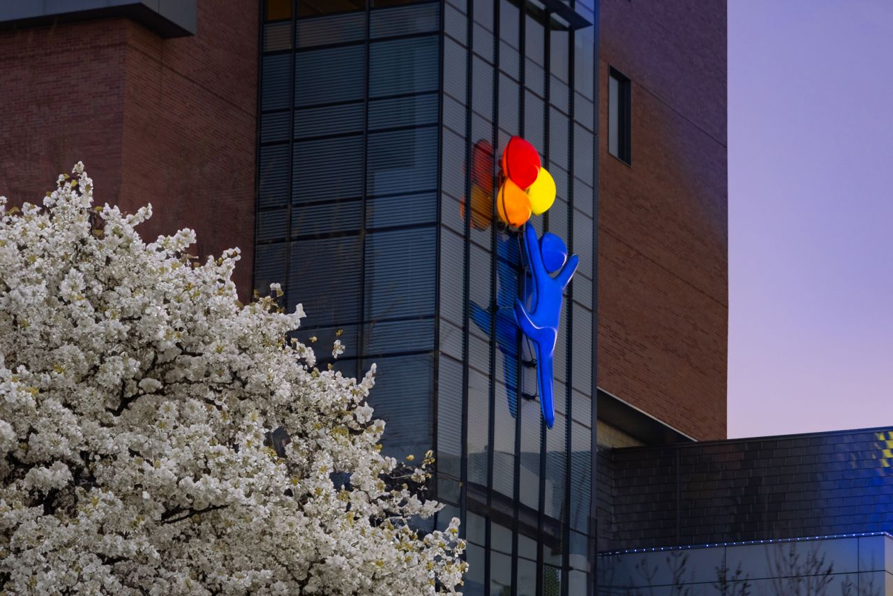 A large, blue sculpture of a person holding colorful balloons is attached to the exterior of a modern brick and glass building, with blooming white flowers visible in the foreground.
