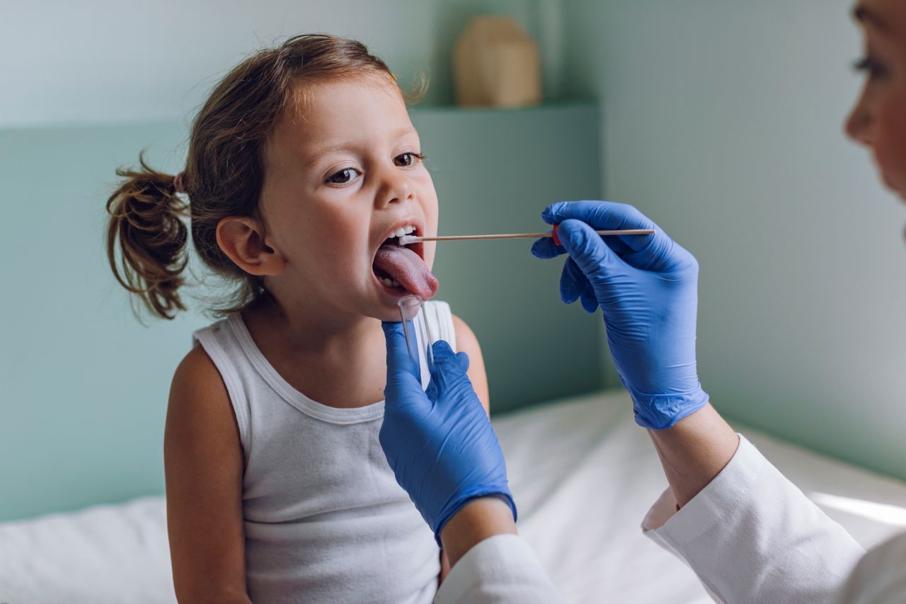 A child with pigtails sits on the table in a doctor’s office. A clinician wearing blue gloves swabs the inside of the child’s mouth with a long cotton swab.