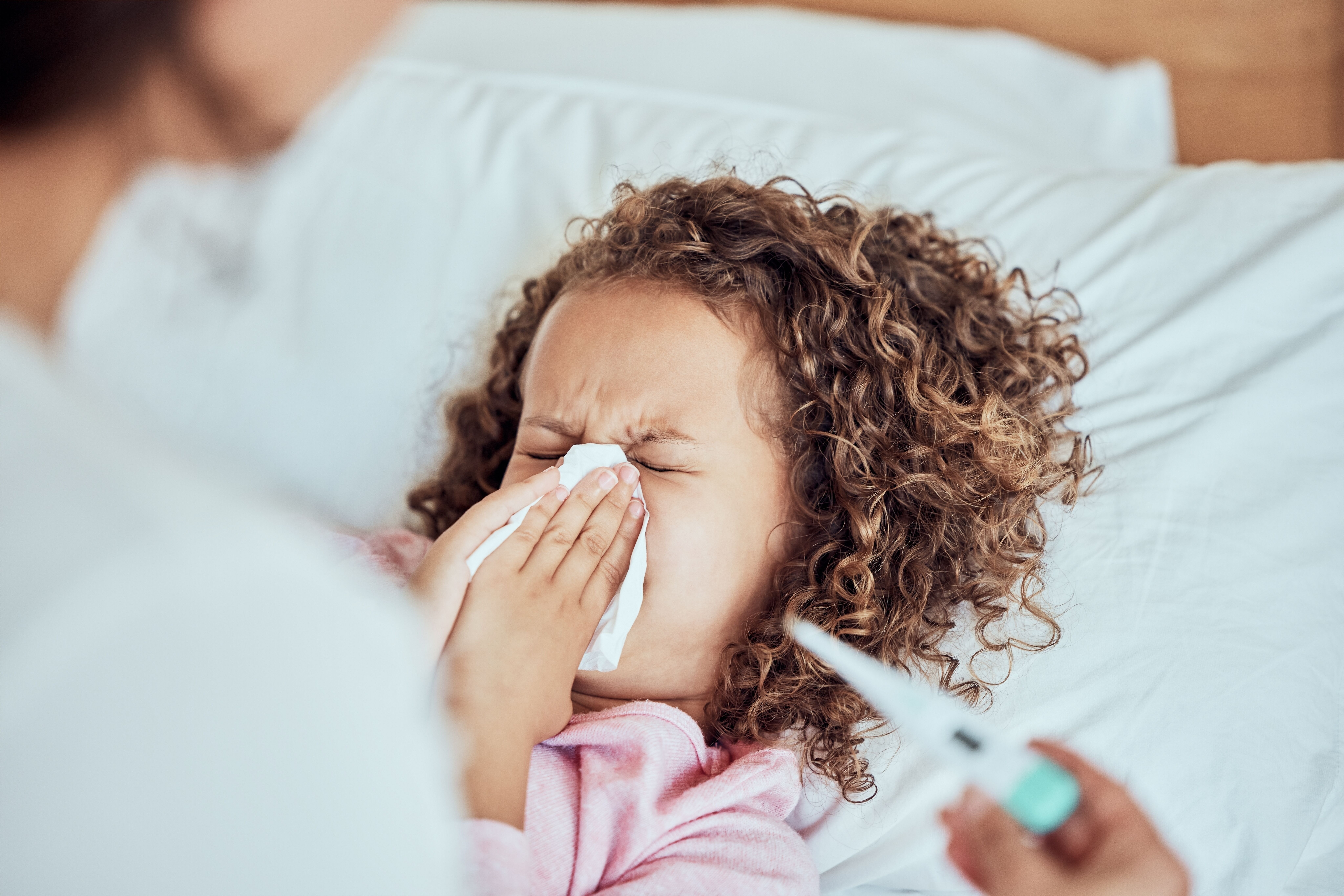 Adult helping child blow their nose while laying in bed with a thermometer 