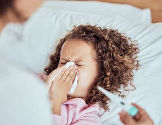 Adult helping child blow their nose while laying in bed with a thermometer 