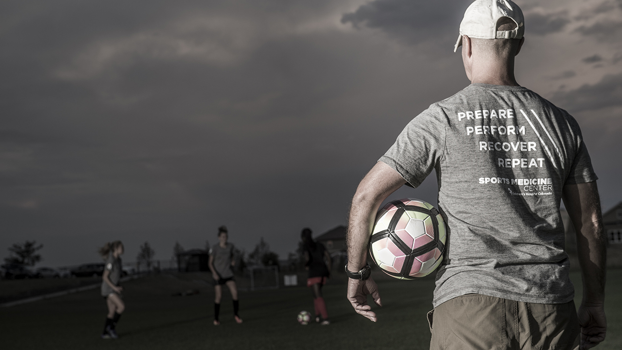 A man with a Children's Hospital sports medicine t-shirt, facing a field, holding a soccer ball