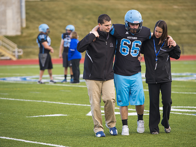 A football player wearing a black jersey, blue shorts and blue helmet is helped off the field by a woman in a black jacket and black pants and a man in a black jacket and khaki pants.