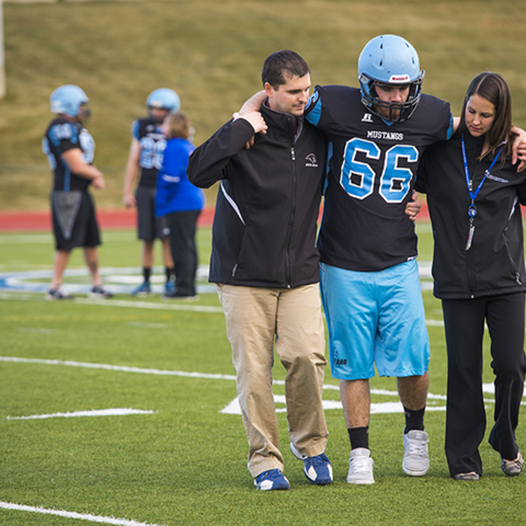 A football player wearing a black jersey, blue shorts and blue helmet is helped off the field by a woman in a black jacket and black pants and a man in a black jacket and khaki pants.