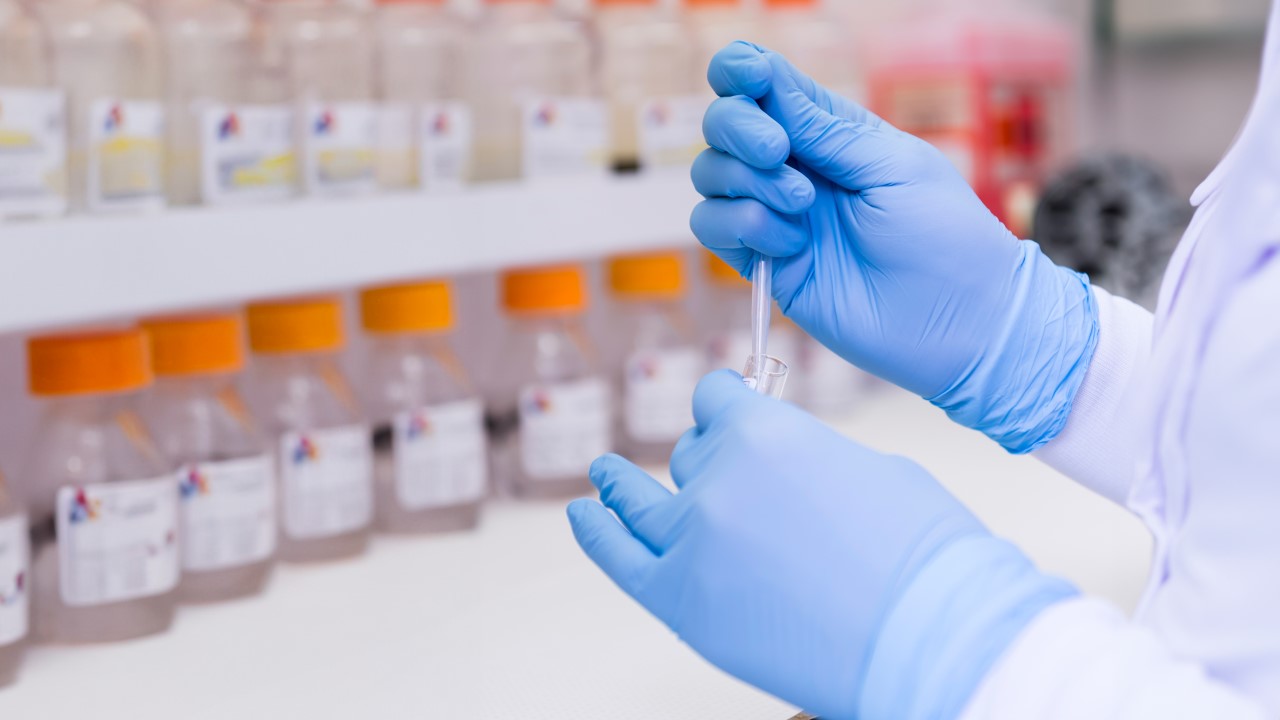 Person in a lab coat wearing blue gloves handles a pipette and test tube in a lab.