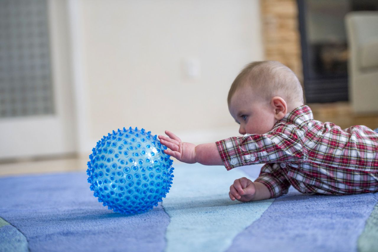 A baby lies on a carpeted floor indoors, reaching one arm toward a round blue textured ball with raised bumps.