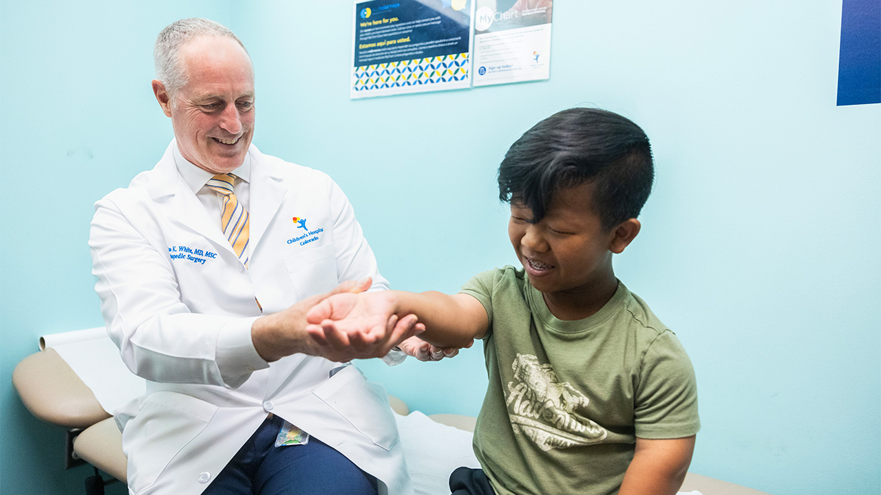 A doctor wearing a white coat smiles and examines a child’s arm.