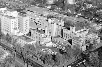 A black and white photo of an aerial view of a city block with a parking lot in the near corner and buildings filling the rest of the block. The building vary in height from three stories to eight stories and all have lots of windows.