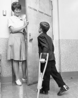 A black and white photo of a women in a white shirt and light color skirt standing in an ajar door greeting a school age boy in dark suit using crutches. The door says Functional Therapy Gymnasium on it.