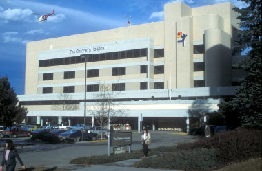 Outside view of The Children's Hospital with three rows of windows and the balloon boy logo on the building while a helicopter flies away in the bright blue sky.