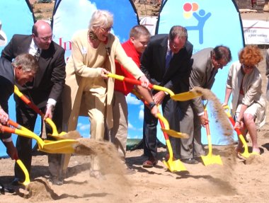 Four men in suits, two women in nice clothes, and a teenage boy in a red polo and khaki pants hold yellow shovels while they dig into the ground in front of Children's Hospital signs