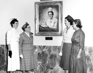 Black and white photo of four women gathered around a photo of a woman on a wall.