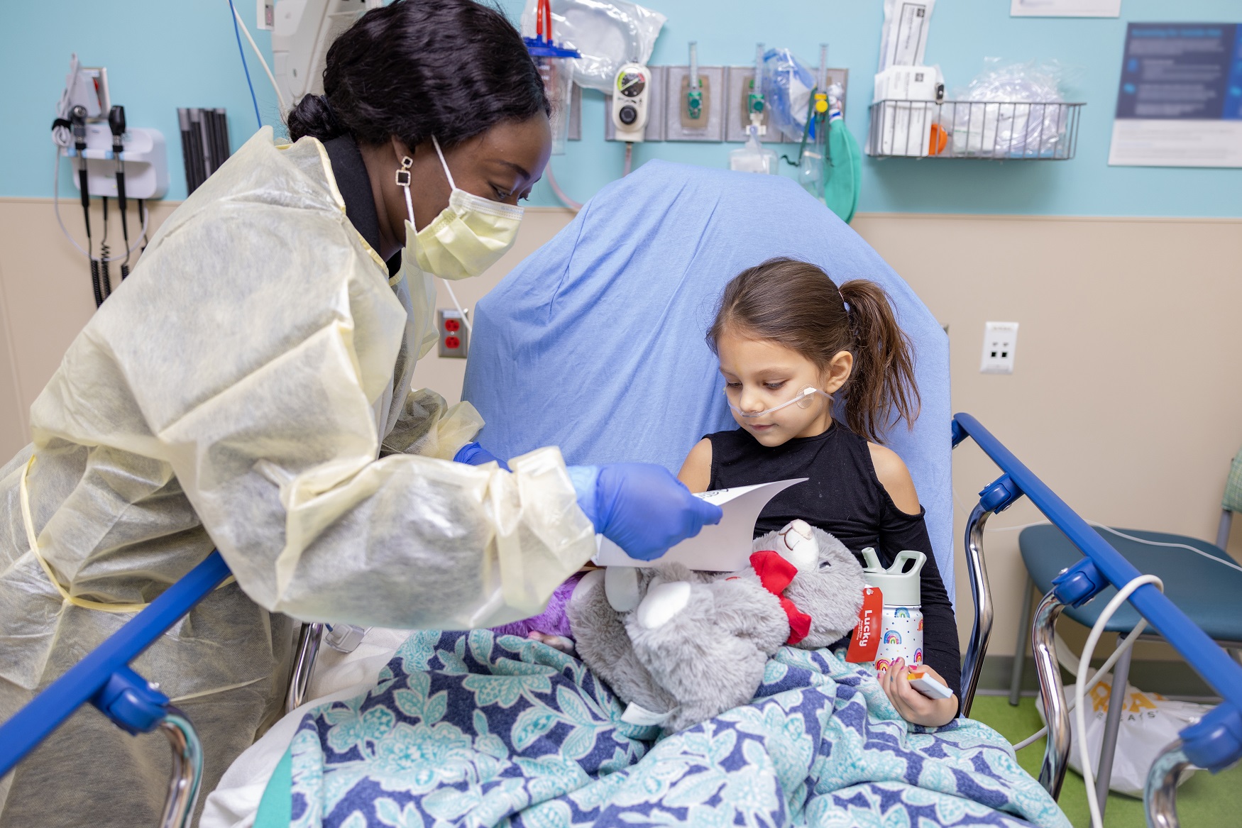 Woman wearing mask and PPE caring for a young girl holding a teddy bear sitting in a pediatric hospital bed. 