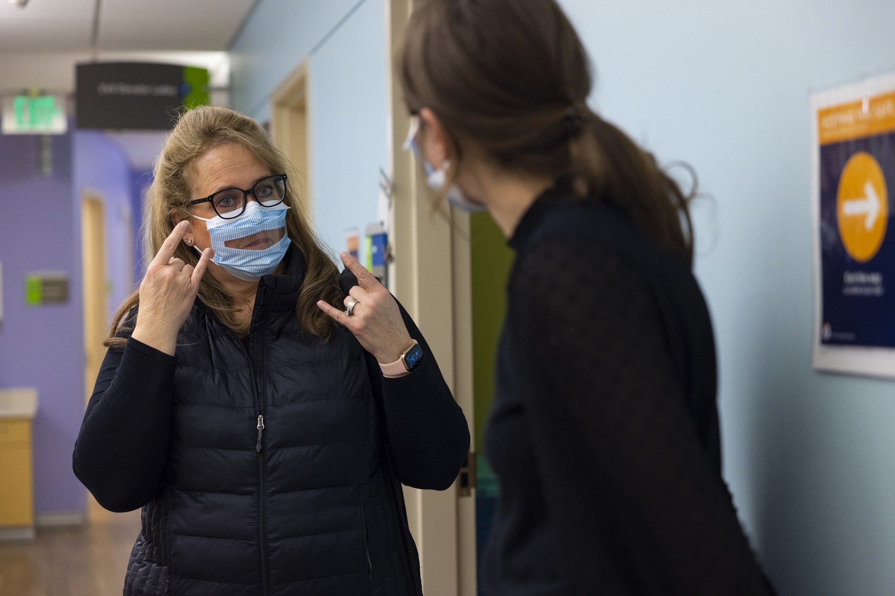 One woman using sign language and wearing a see-through mask, communicating with another woman. 