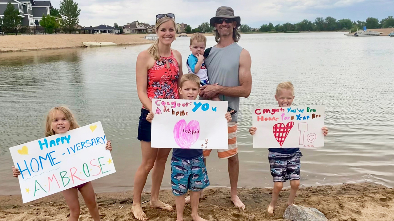 Ambrose's family in front of a lake. The three other children are holding signs congratulating Ambrose on the anniversary of his coming home from the hospital.