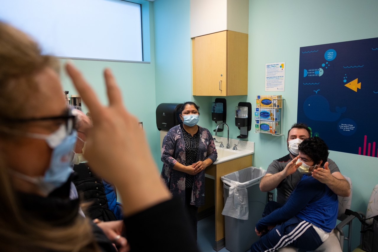 A medical interpreter speaks with Juan in sign language as Angel sits on his lap. Maria stands next to them as she listens to the providers speak.