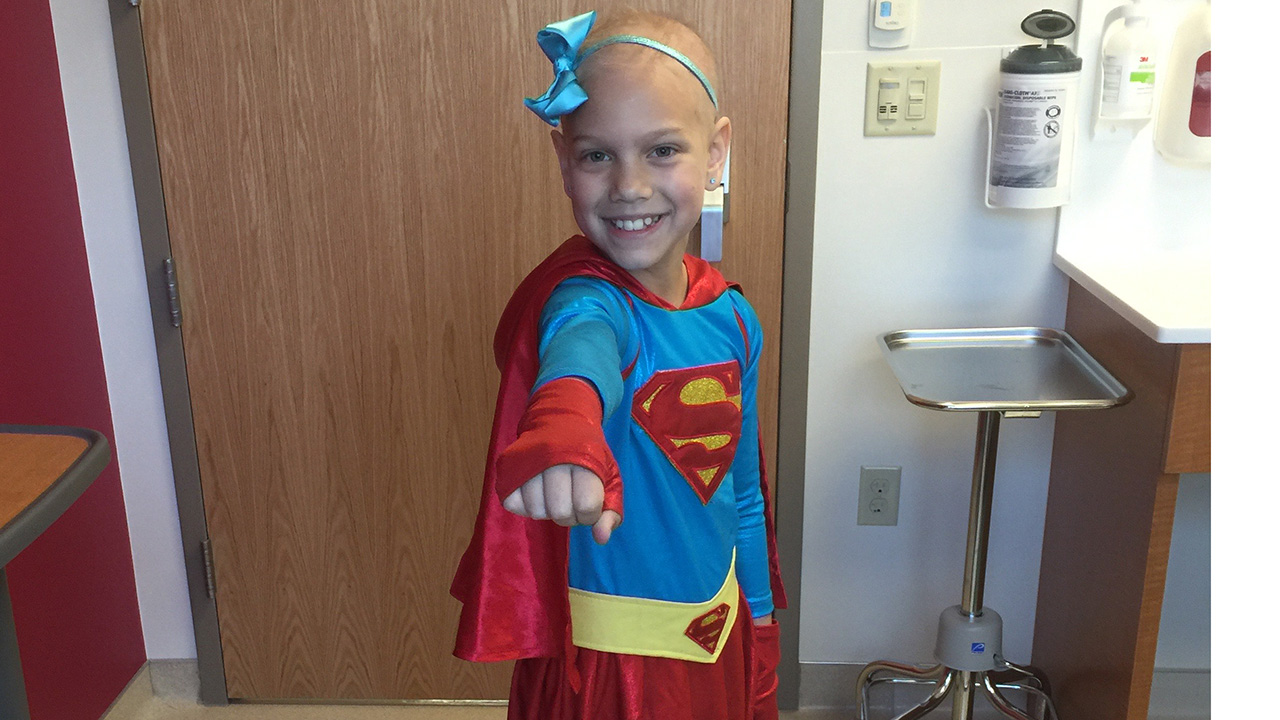 A child dressed in a Superman costume, smiling and posing confidently in a medical office.