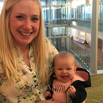 Young blond mother holding baby girl facing forward. Baby looks as if she's giggling; mom is smiling.