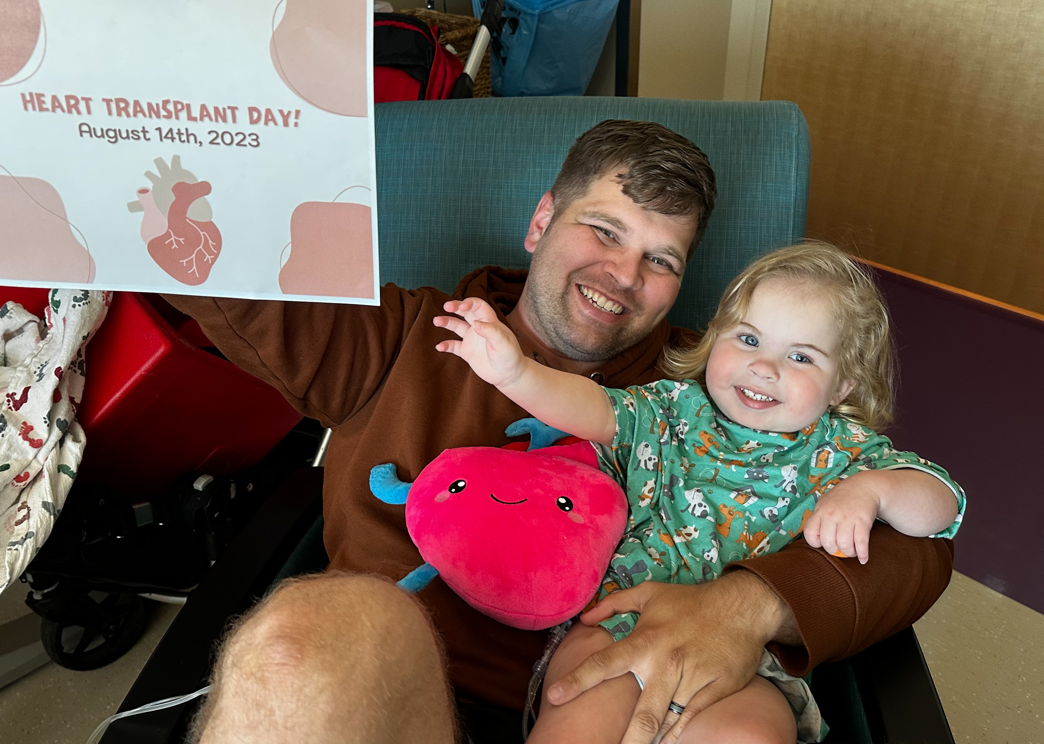 Willow sits on her father's lap at the hospital while he holds a sign that says, "Heart Transplant Day! August 14th, 2023"