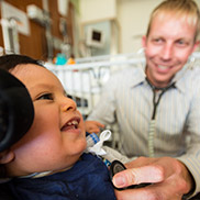 A doctor with blond hair and a white striped shirt listens to a young boy's heart through a stethoscope.