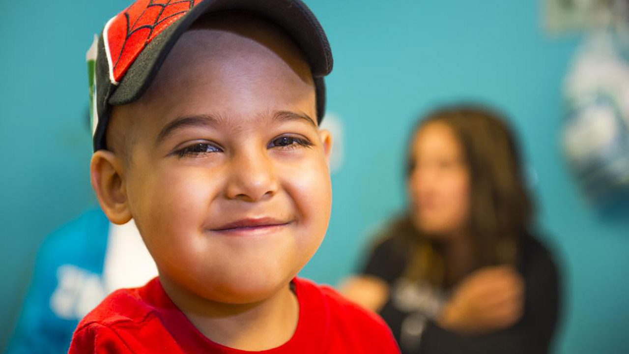 A boy with a red spider hat and red shirt that says SLUGGER 8 on it smiles in the foreground while two adults talk to a doctor in the background.