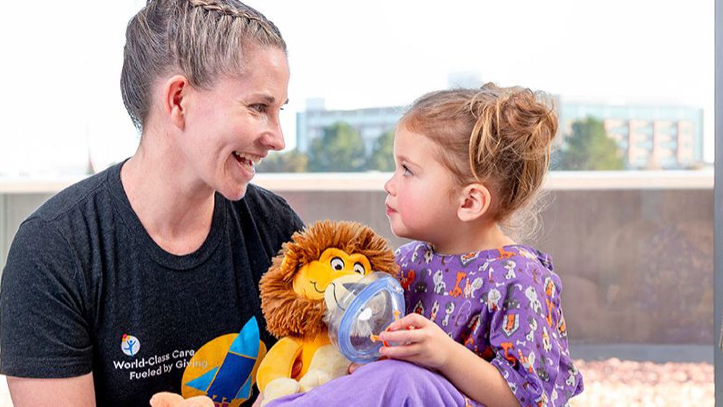 A child life specialist helps a kid prepare for a medical procedure by using a breathing mask on a stuffed animal lion.