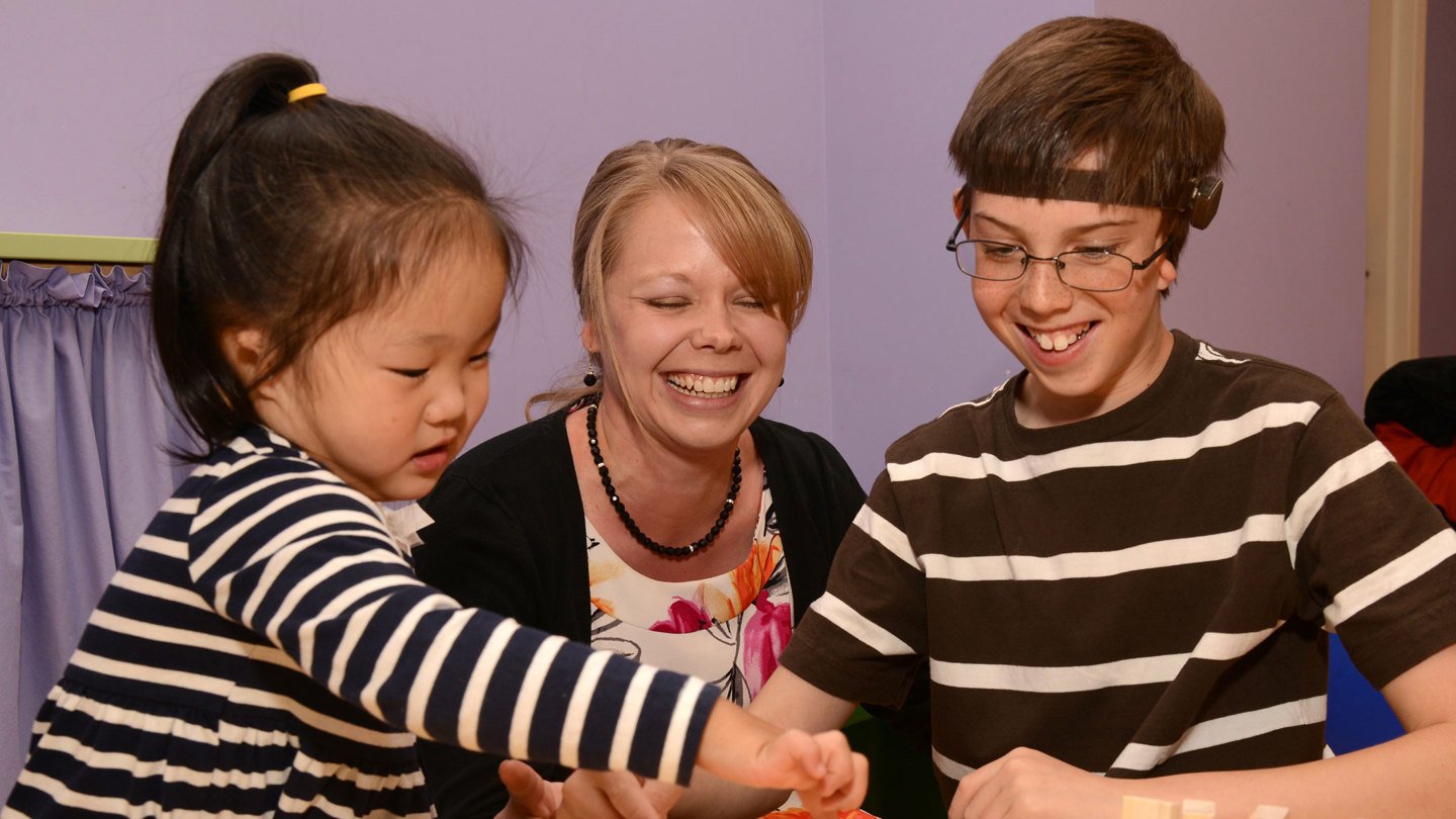 Audiologist Dr. Hedman sits in the middle of two patients playing Jenga. The girl on the left as her black hair in a ponytail and is wearing a black and white striped shirt. The boy on the right has glasses and is wearing a brown shirt with white stripes.