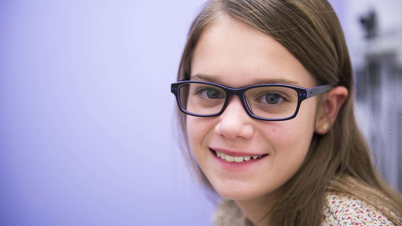 A girl with long brown hair and glasses smiles at the camera.