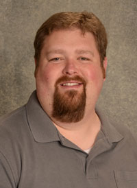 A headshot of heart perfusionist Cory Ellis who has light brown hair and a goatee and is wearing a gray polo.