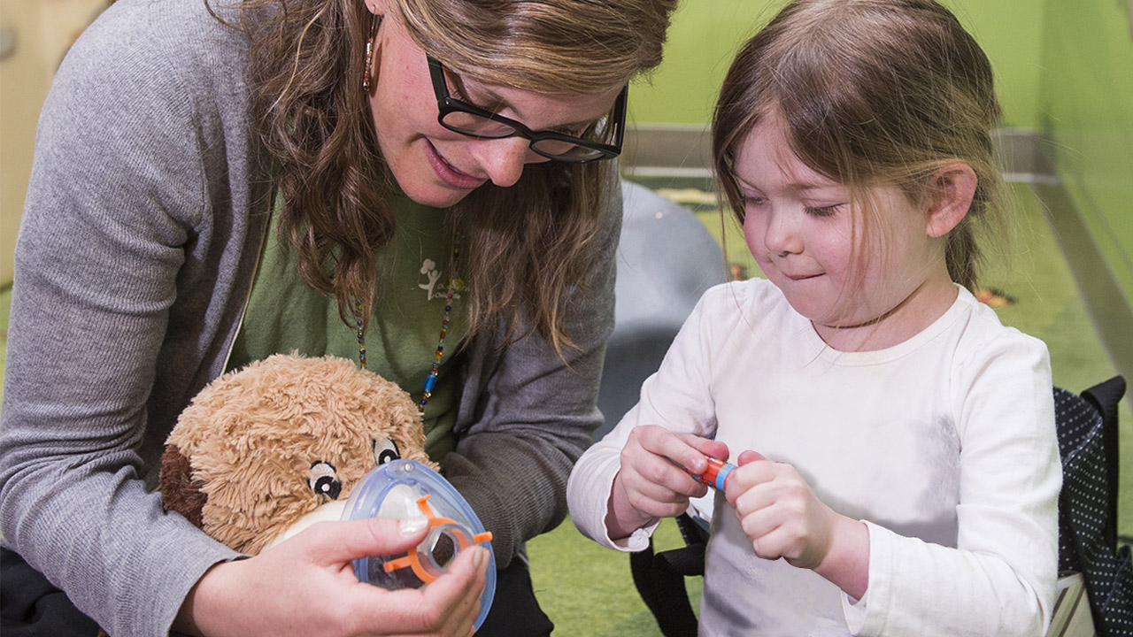 A child life specialist teaches a girl about anesthesia by placing a mask on a stuffed animal