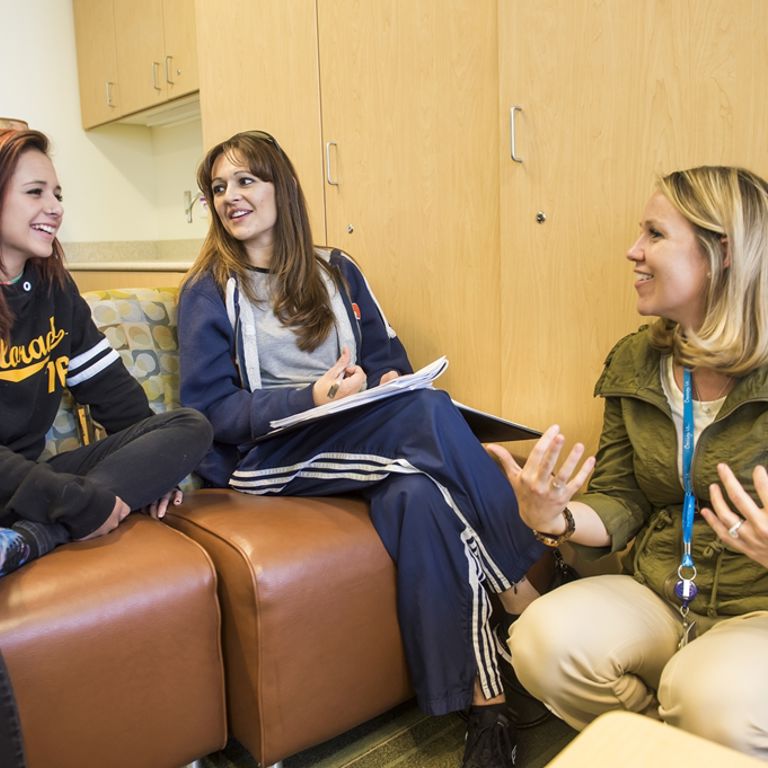 A blonde woman wearing yellow pants and a green jacket squats next to two ladies sitting in brown chairs while they have a conversation. They both have long brown hair. One is wearing a black sweatshirt with Colorado in yellow lettering and the other is wearing a blue track suit and taking notes in a notebook.