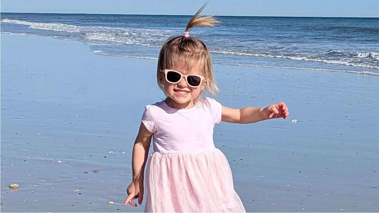 A toddler girl wearing sunglasses and smiling on the beach. She received care for fetal heart block at Children’s Colorado.