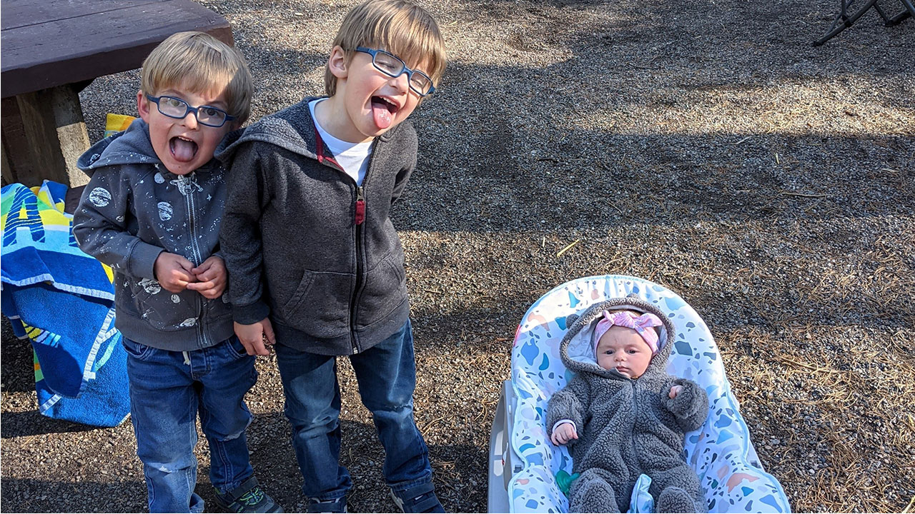 A baby girl sits in a rocker at a campsite while her two older brothers make silly faces while standing next to her.