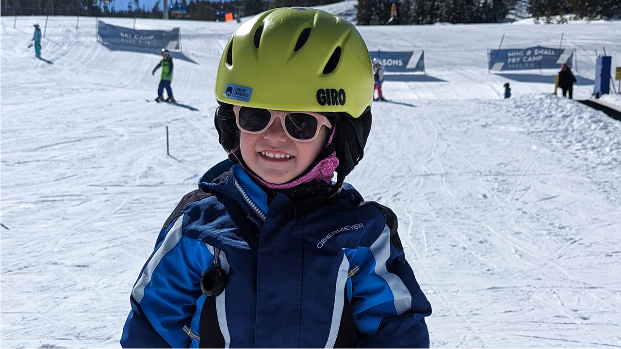 A toddler girl smiles on a ski slope while wearing a yellow helmet and blue jacket.