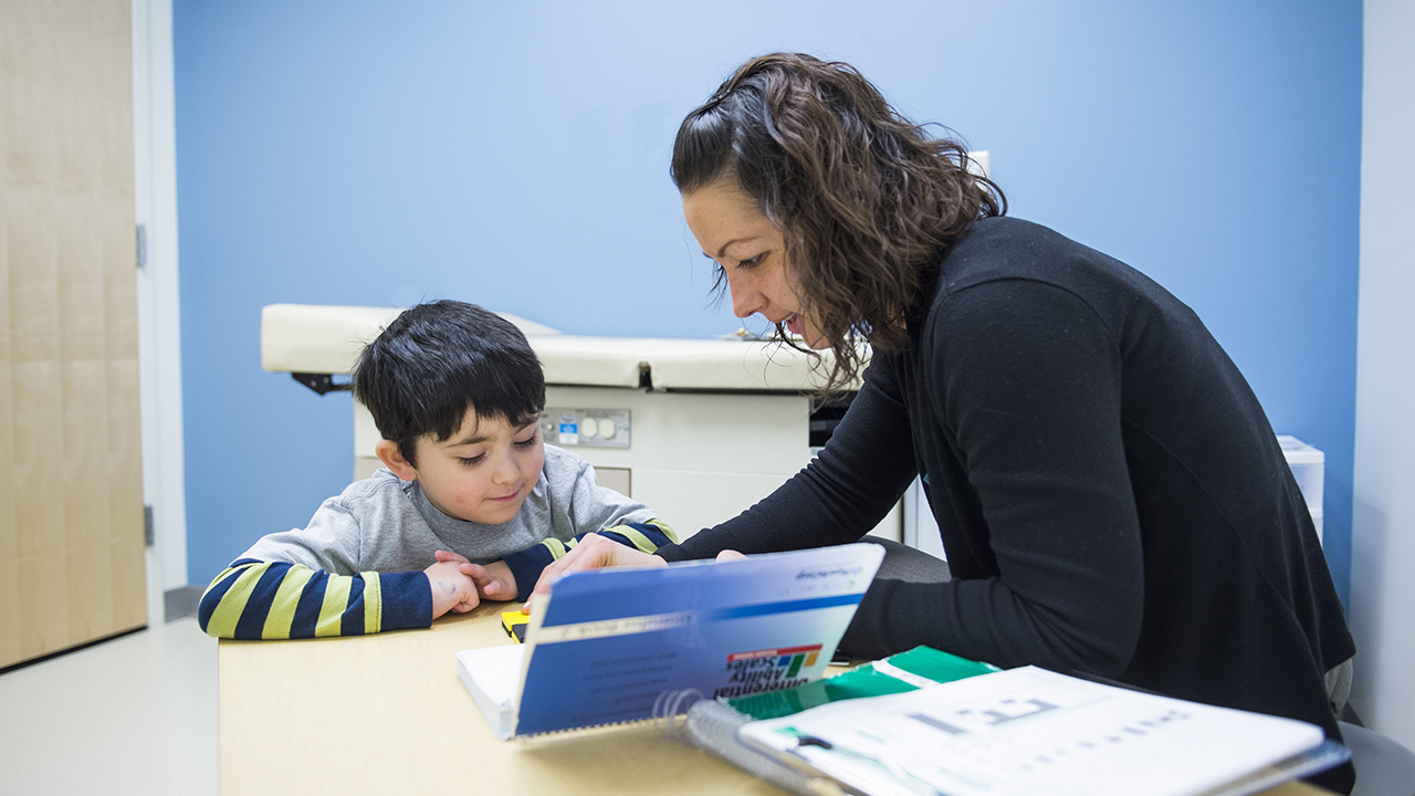 A boy wearing a blue and yellow striped shirt with a gray short sleeve shirt over it listens to a woman with short brown hair and wearing a black cardigan.