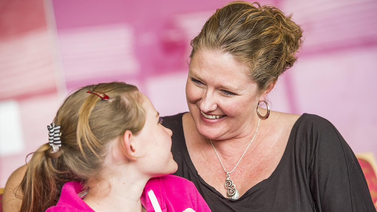 A close-up of a woman in a black shirt and a girl in a pink sweatshirt smiling at each other.