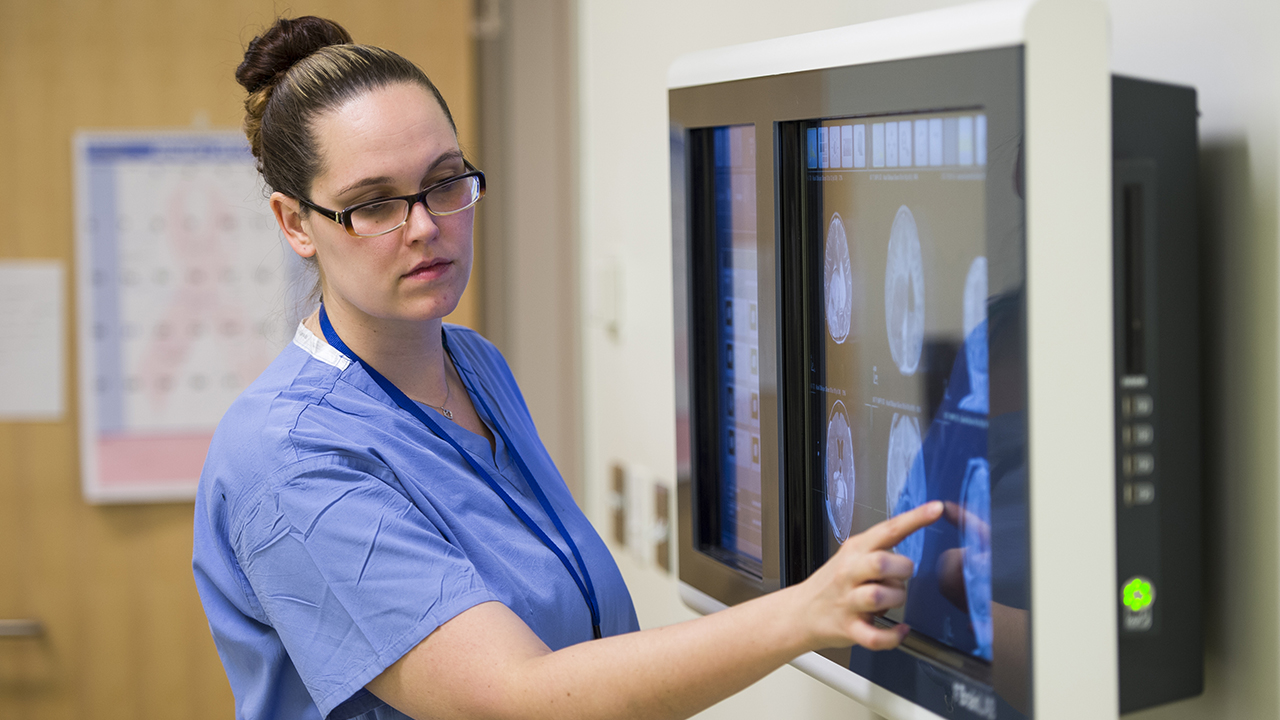 A woman in blue scrubs and glasses with her hair in a bun points to a screen with brain images on it.