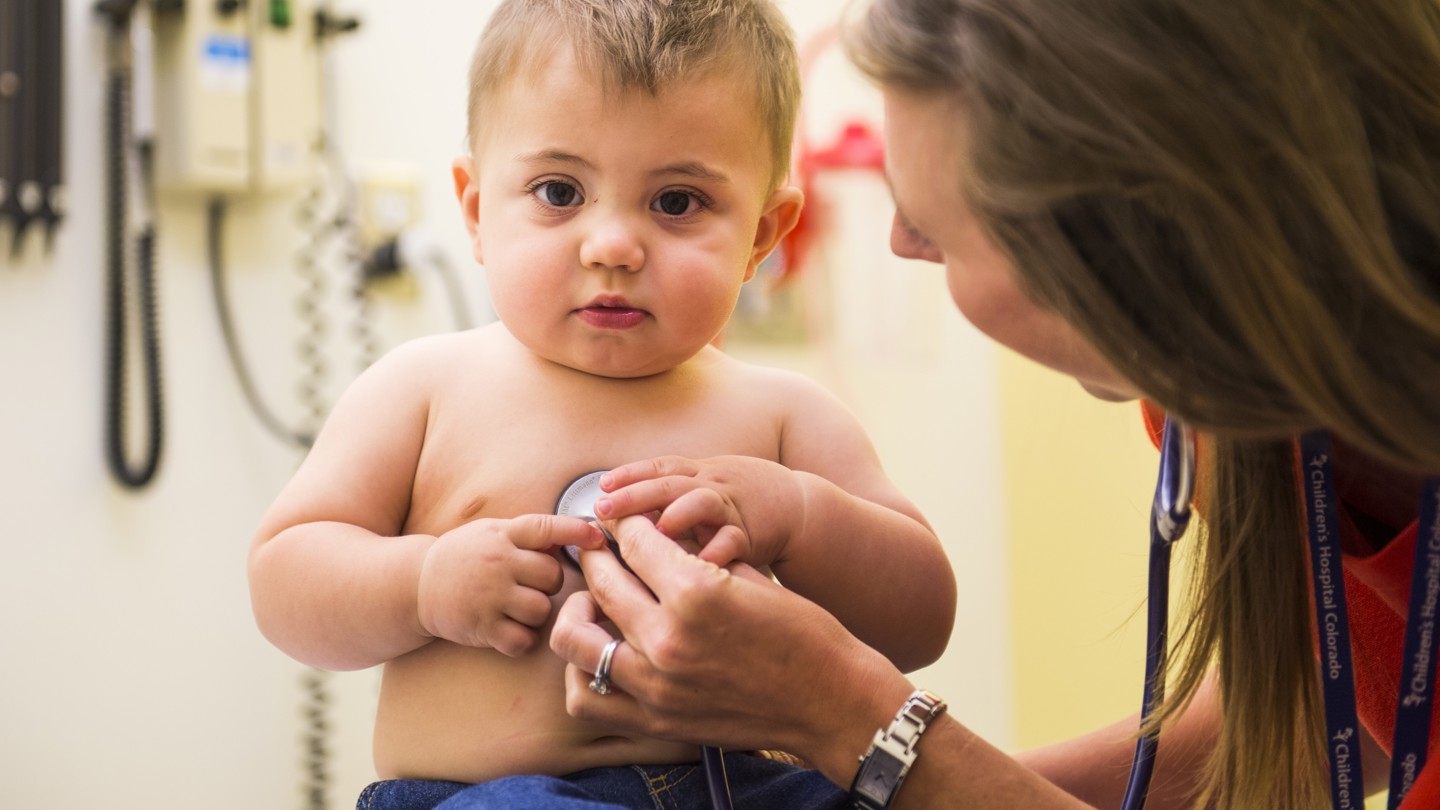 A toddler with brown hair sits in a doctor's office while a doctor examines him with a stethoscope.