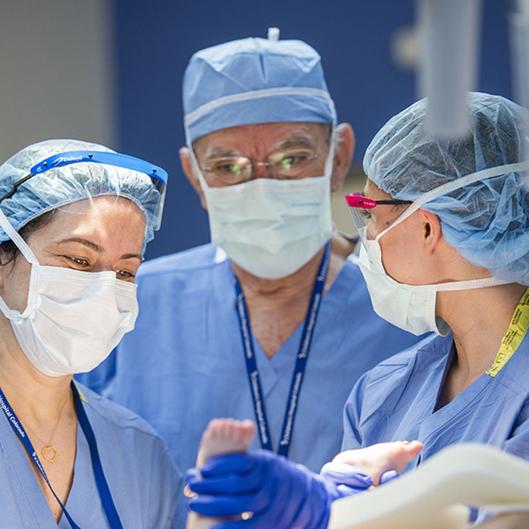 Dr. Alberto Pena, wearing light blue scrubs, a hair covering and white mask stands at an operating table with two women in matching light blue scrubs holding a baby's feet with blue latex gloves.