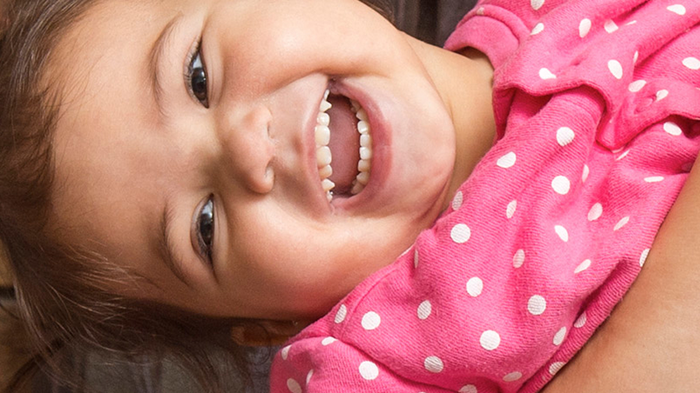 A young toddler with brown hair and dressed in a pink cardigan with white polkadots is lying down and laughing.