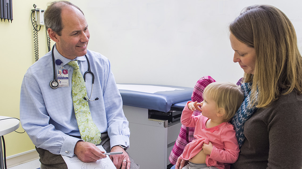 A doctor wearing a blue shirt and green tie points to a toddler's belly while she sits on her mom's lap.