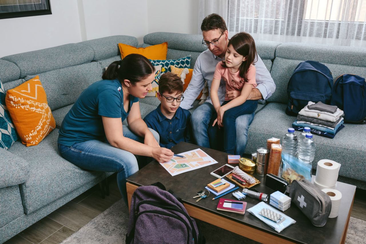 A mother, father, son and daughter gather around their coffee table while discussing a emergency plan on a map of their house. Emergency items like canned food and a first aid kit sit on the table in front of them.