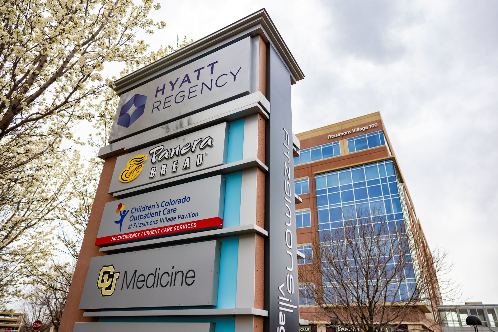 Tall outdoor sign listing ‘Hyatt Regency,’ ‘Panera Bread,’ and ‘Children’s Colorado Outpatient Care Pavilion – Urgent Care Services,’ with a CU Medicine logo, standing in front of a multi-story glass-and-brick building and trees.