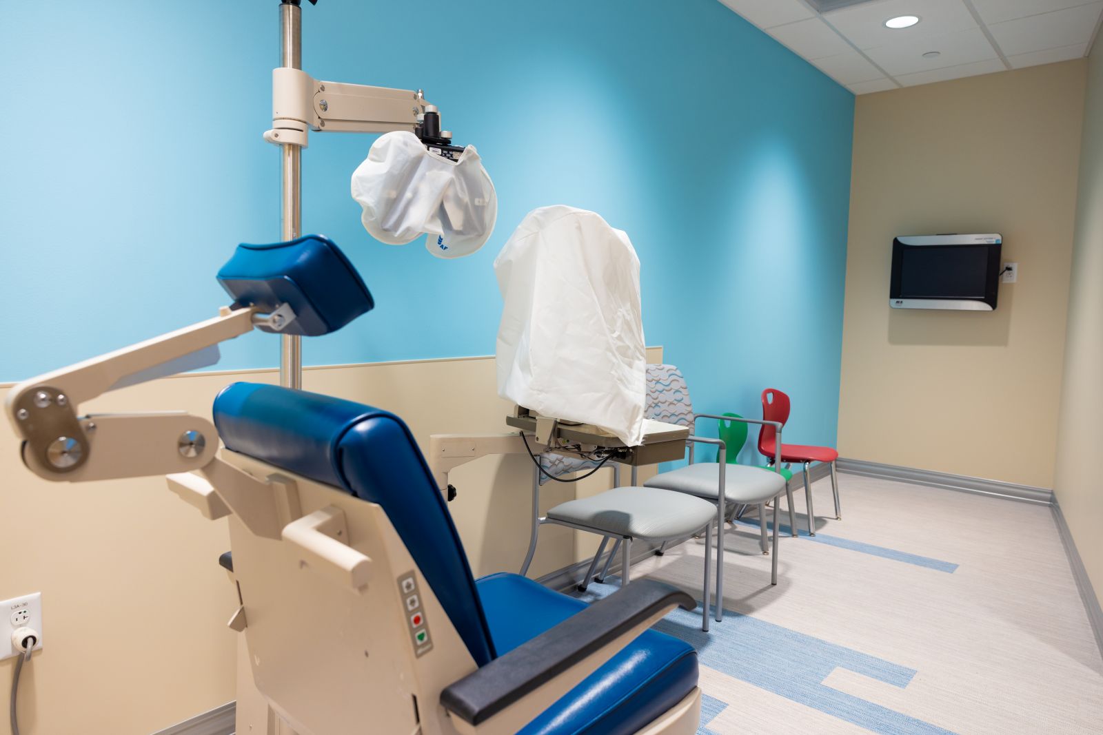 Blue medical examination chair with attached adjustable light and armrests in a clean exam room, with additional chairs, a wall-mounted screen, and blue and beige walls.