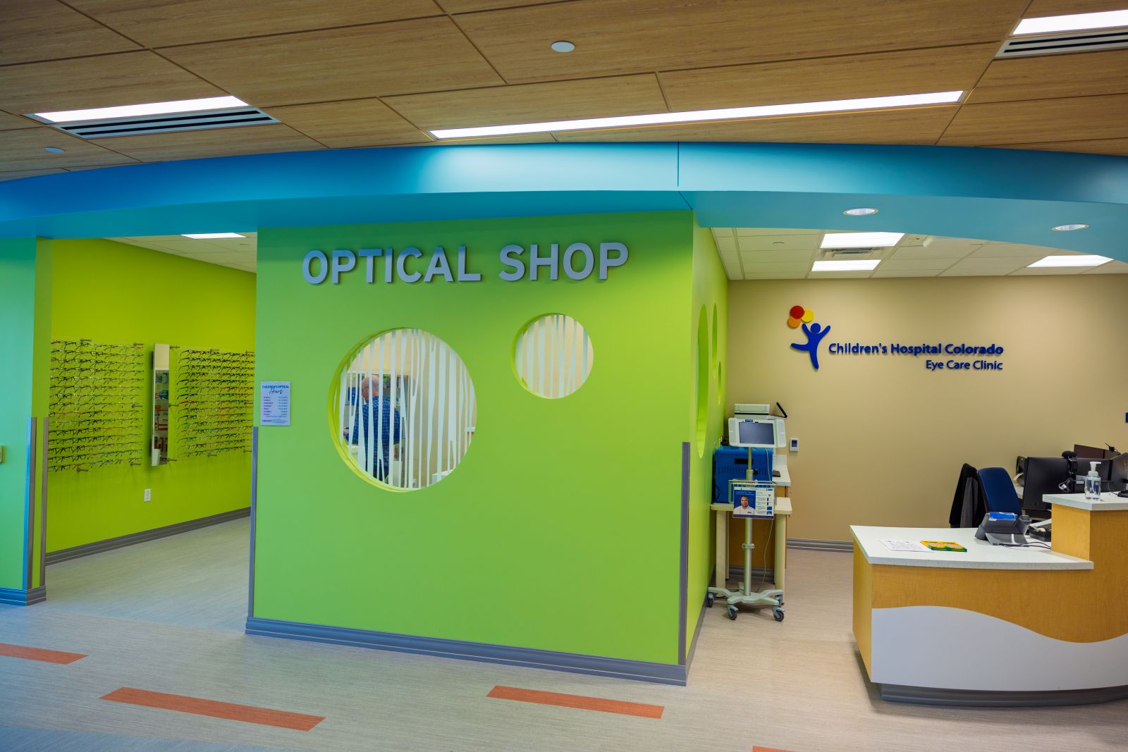 Bright clinic reception area with a green wall labeled ‘Optical Shop,’ adjacent to a check-in desk labeled ‘Children’s Hospital Colorado Eye Care Clinic.'