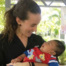 A woman with long brown hair holds a baby wearing a red a blue shirt.