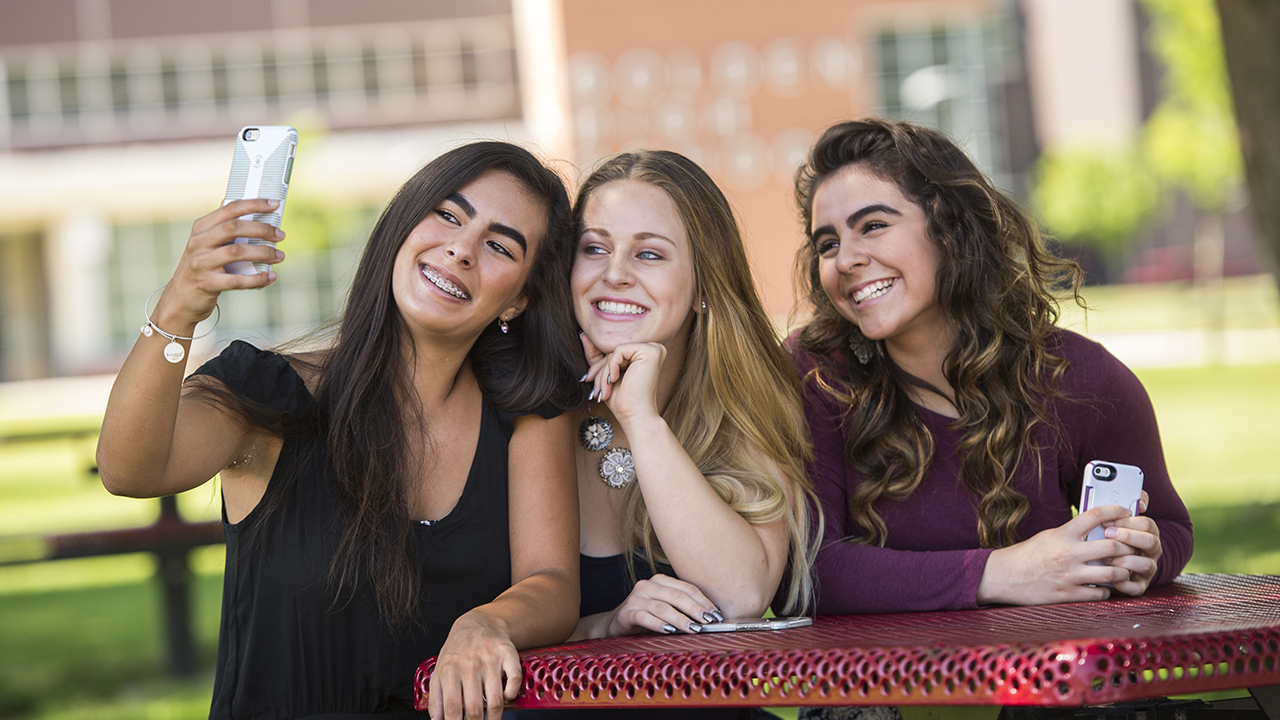 Three teenage girls take a group selfie. The girl on the left with long, dark brown hair is holding the camera phone and wearing a black short sleeve blouse. Next to her is a girl with long, light brown hair who is resting her head on her hand. The girl on the right has long, brown curly hair and is wearing a plum sweater.