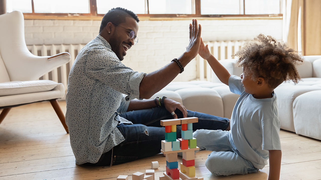 A dad and school-aged child sit on the floor smiling and high-fiving. A tower of colorful blocks is between them.