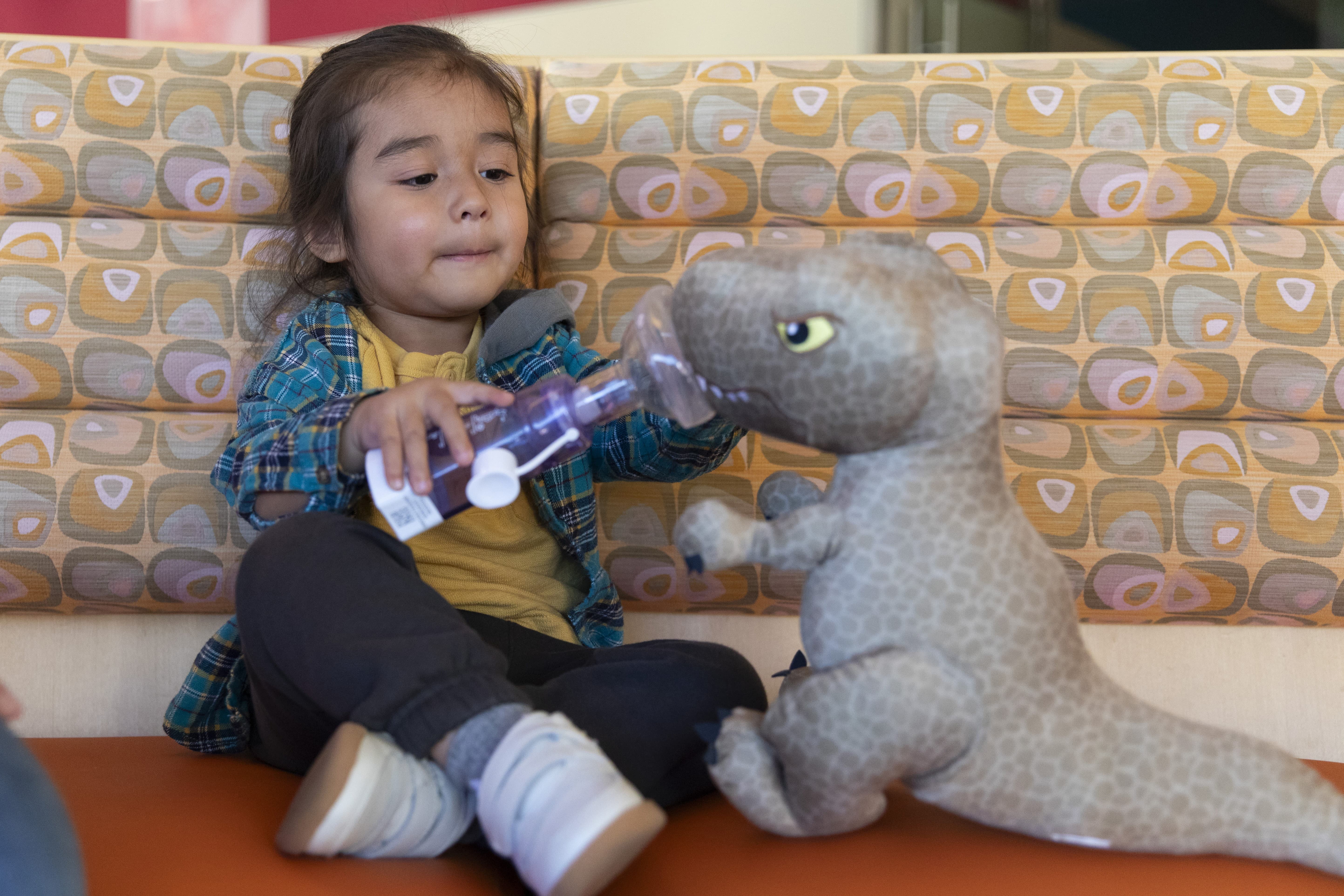 A young child pretends to use an inhaler on a stuffed toy dinosaur. 