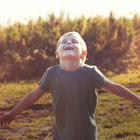 A school-age boy wearing a short sleeve gray shirt stands in a field with tall grasses behind him while the sun shines down. He is looking up smiling with his eyes closed and his arms out to the side.