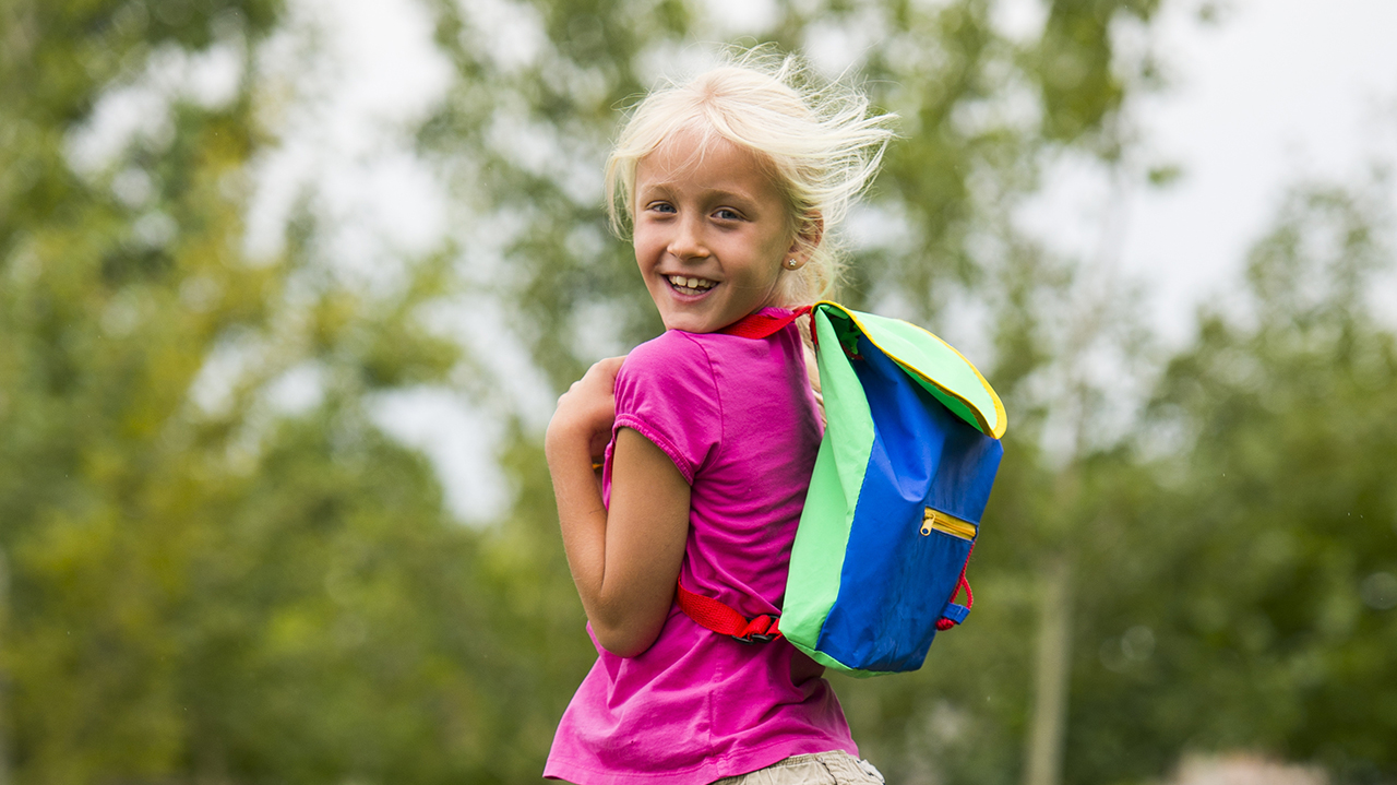 A girl with blonde hair and wearing a pink t-shirt and blue backpack stands in front of green trees.